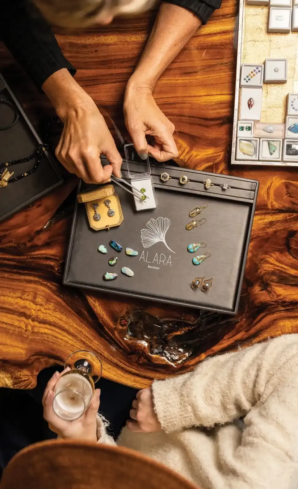 Not Just Jewelry 3 Overhead view of hands at a wooden table with a tray of loose gemstones and jewelry