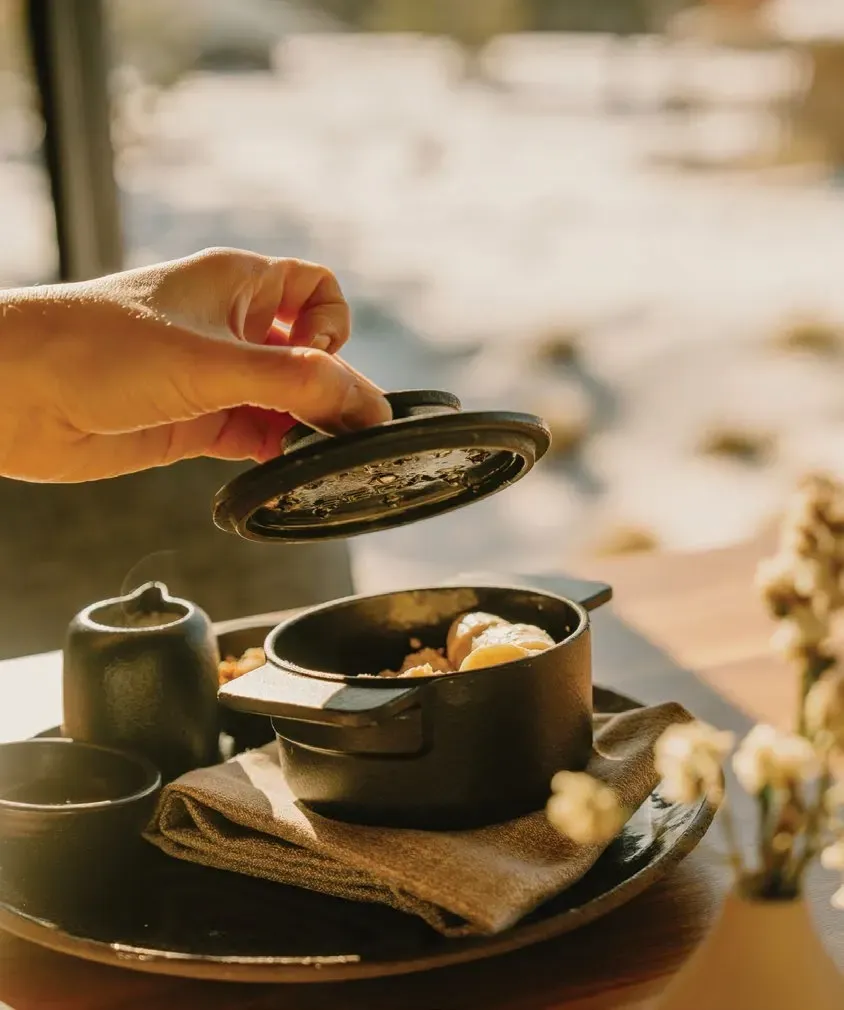A hand lifts the lid from a small cast iron pot beside a snowy window