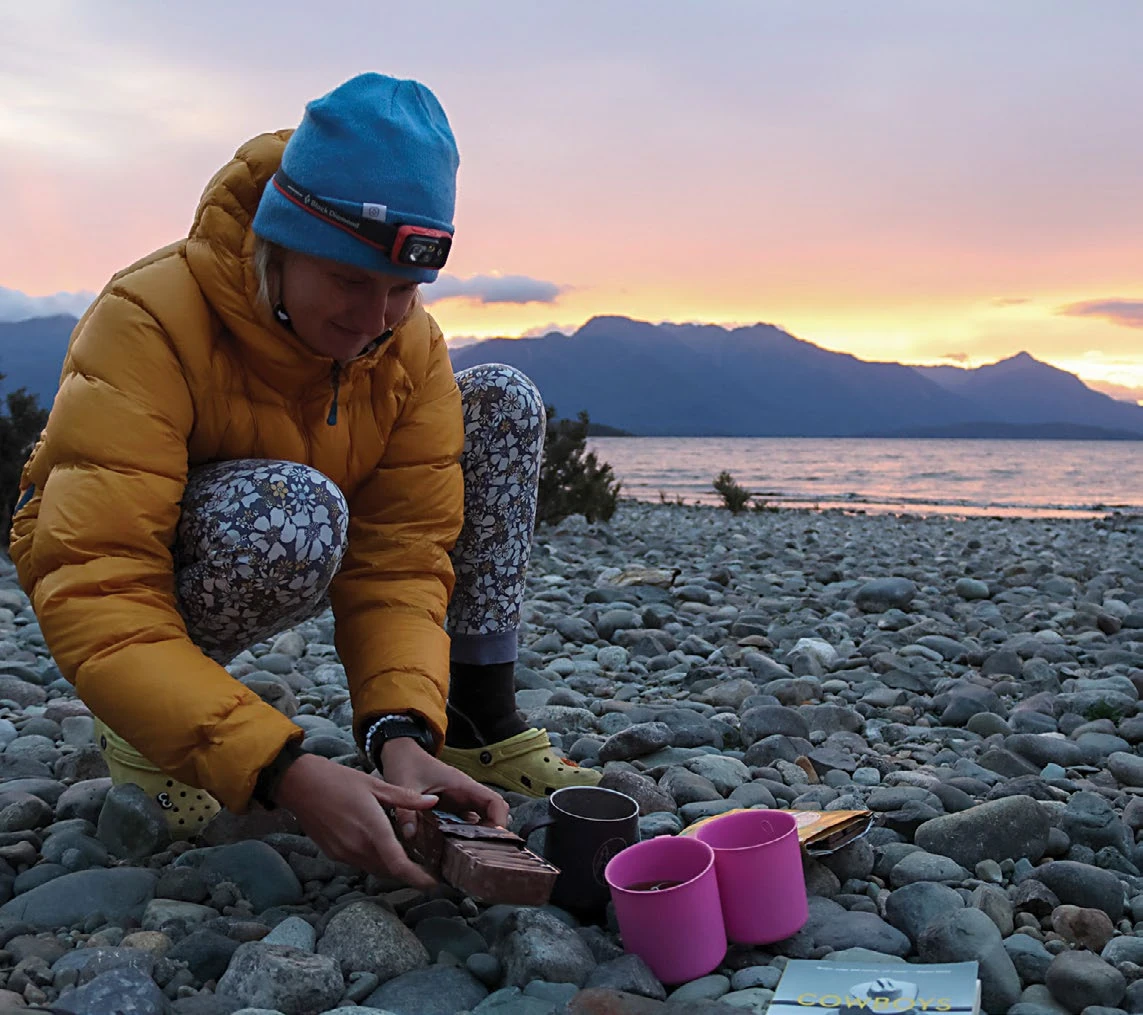 A camper in a yellow puffer jacket dips a Tim Tam at sunset on a rocky beach