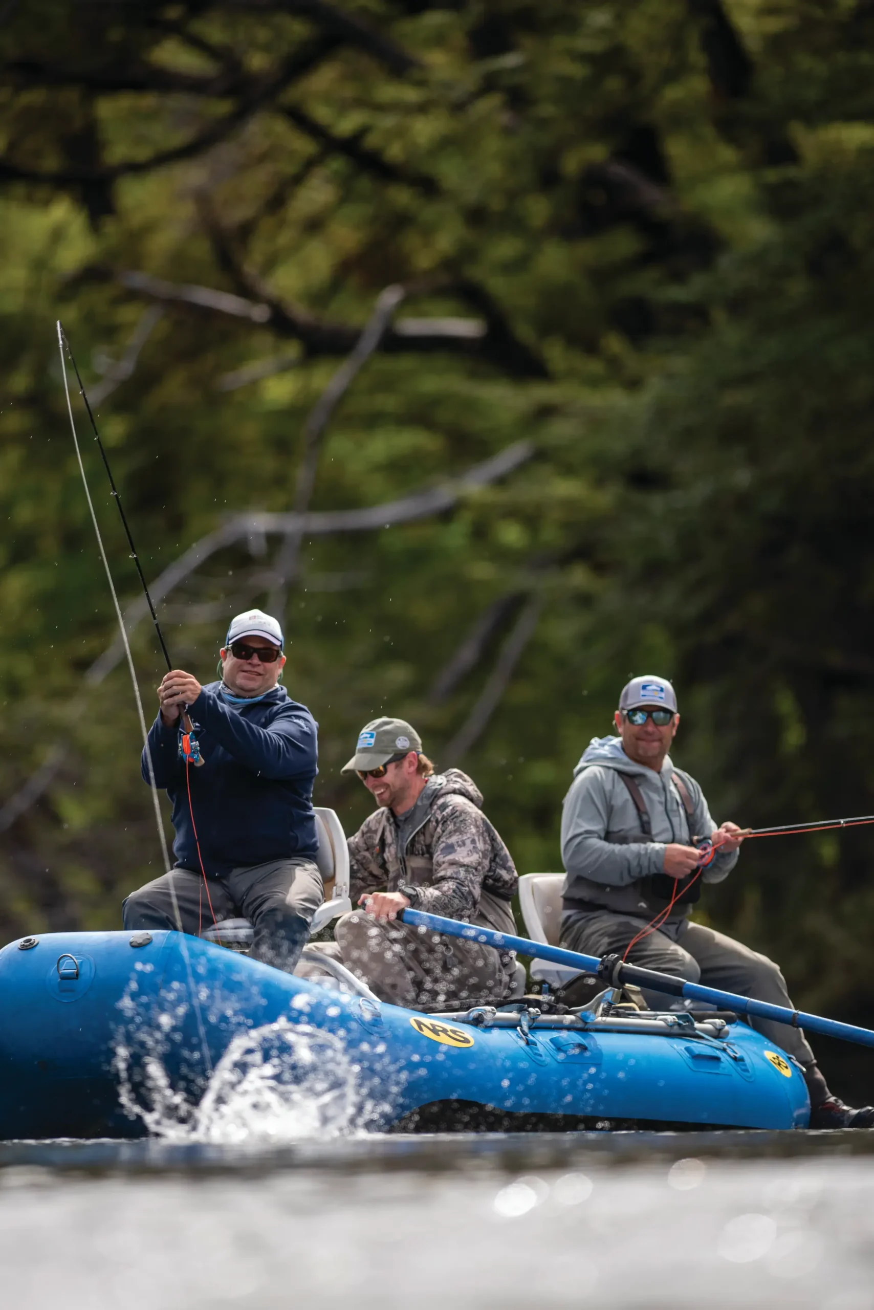 Three anglers fly fishing from a blue raft, one hooked up, against dark forest